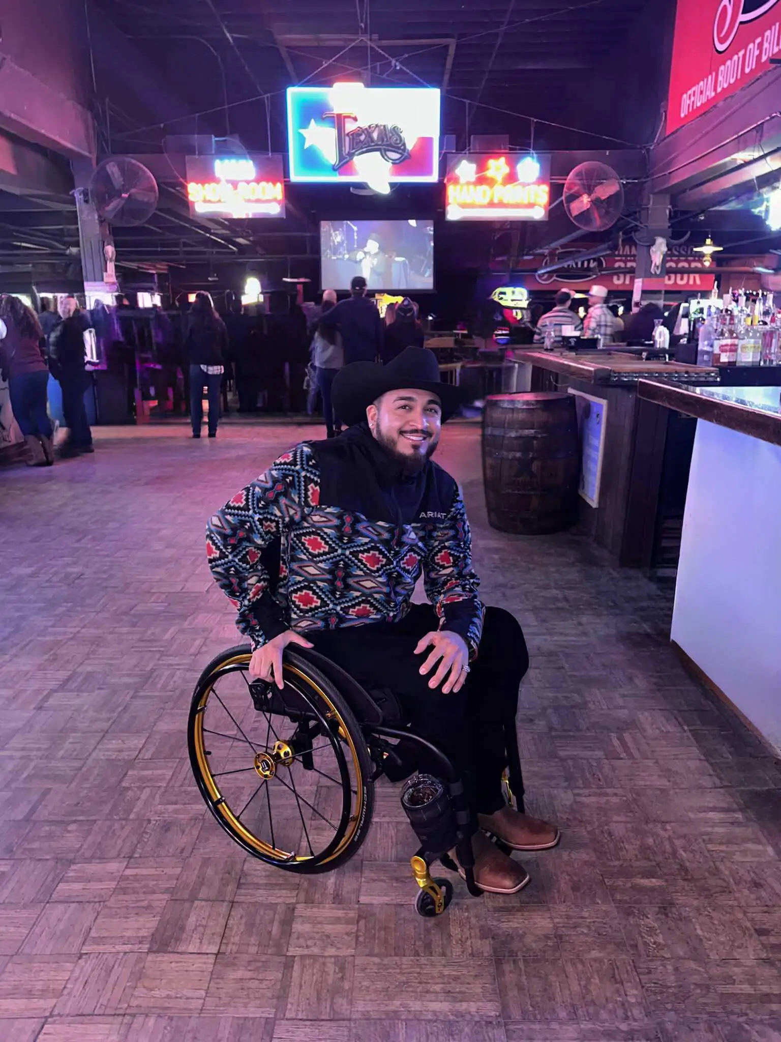 A picture of Anthony Sanchez out on the town, in the famous Fort Worth venue known as Billy Bobs. Dressed in a black cowboy hat, a black puffy jacket, and black jeans with holes over the knee, he's ready to show that being disabled doesn't mean you can't live a good life. Also, in the background, various fixtures of Billy Bobs can be seen out of focus, including a bar and the neon sign of the logo.
