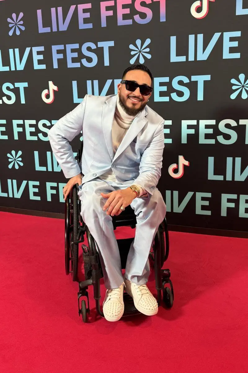 A picture of Anthony Sanchez out on the town, in the famous Fort Worth venue known as Billy Bobs. Dressed in a black cowboy hat, a black puffy jacket, and black jeans with holes over the knee, he's ready to show that being disabled doesn't mean you can't live a good life. Also, in the background, various fixtures of Billy Bobs can be seen out of focus, including a bar and the neon sign of the logo.